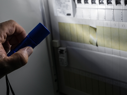 A hand holds a flashlight to look at an electrical panel in the dark.